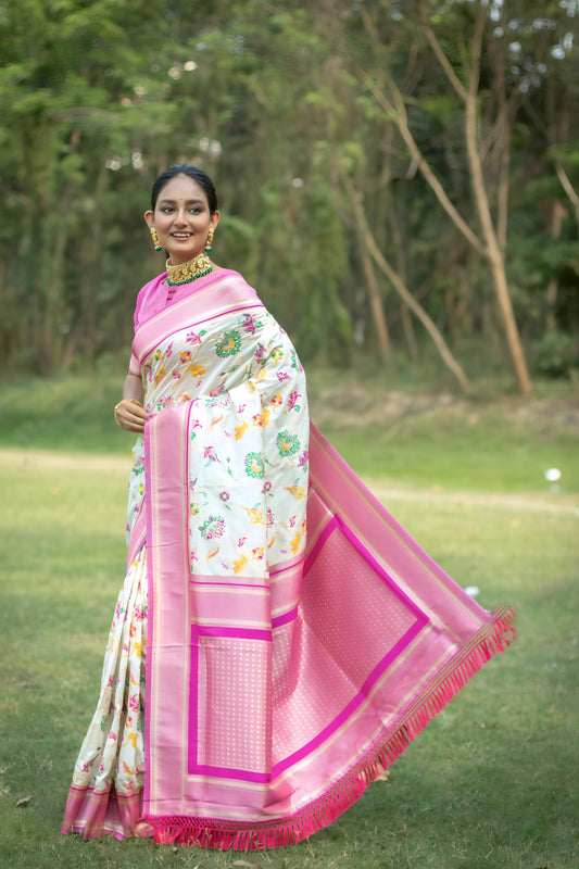 Woman wearing an off-white silk saree with pink borders and multicolor floral motifs, paired with a pink blouse and traditional gold jewelry, elegantly standing on a green lawn with trees in the background.