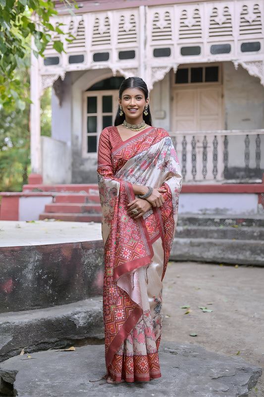 Woman wearing an off-white silk saree with red Patola-inspired geometric and floral borders, paired with a matching blouse and traditional jewelry, elegantly posing on stone steps in front of a vintage heritage house with ornate wooden railings and lush green trees in the background.