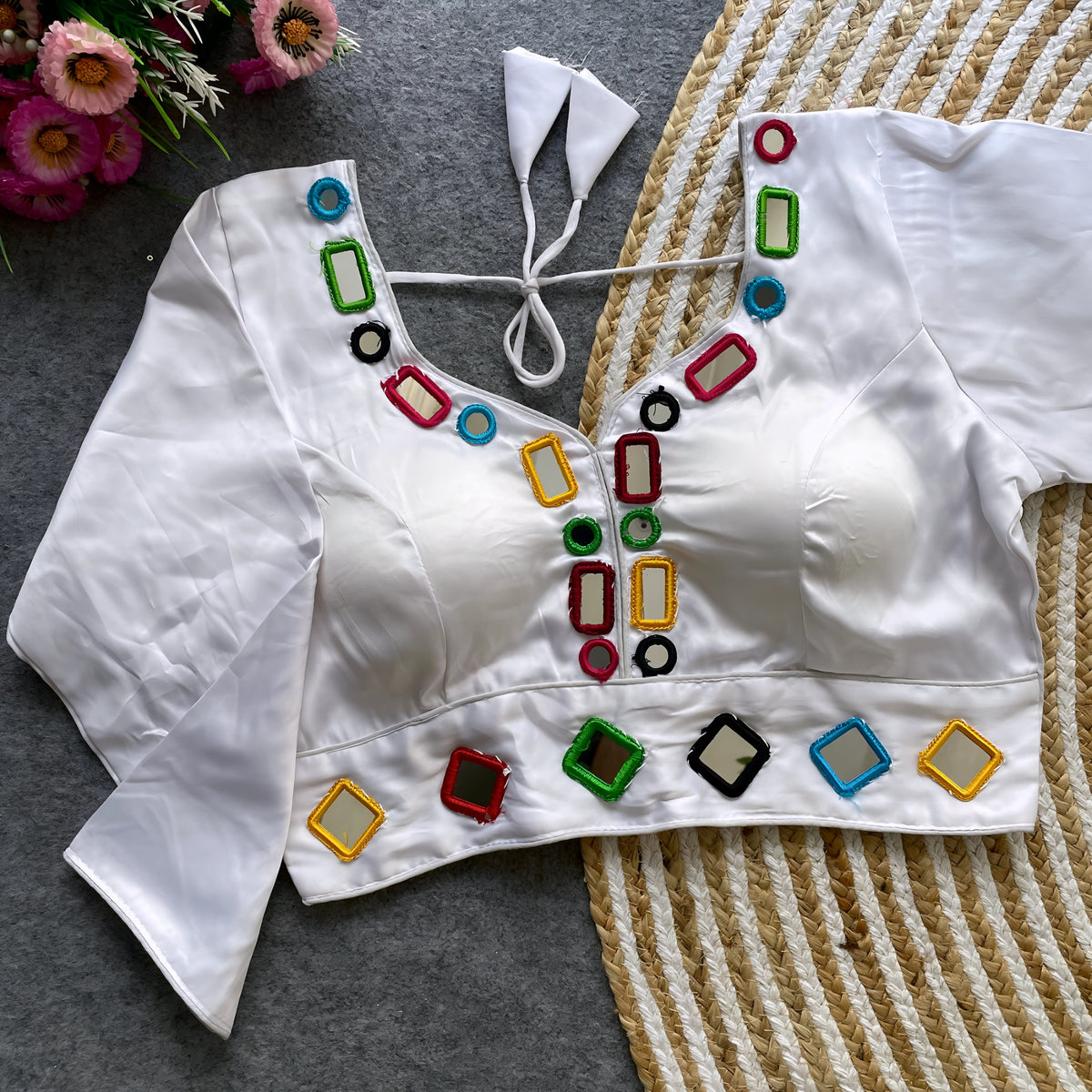 Off-white blouse embellished with square and round mirror work featuring multicolored embroidered borders, stylish back tie-up details, and three-quarter sleeves. Displayed flat on a textured grey surface with a woven mat and pink flowers, highlighting its festive and contemporary ethnic design.
