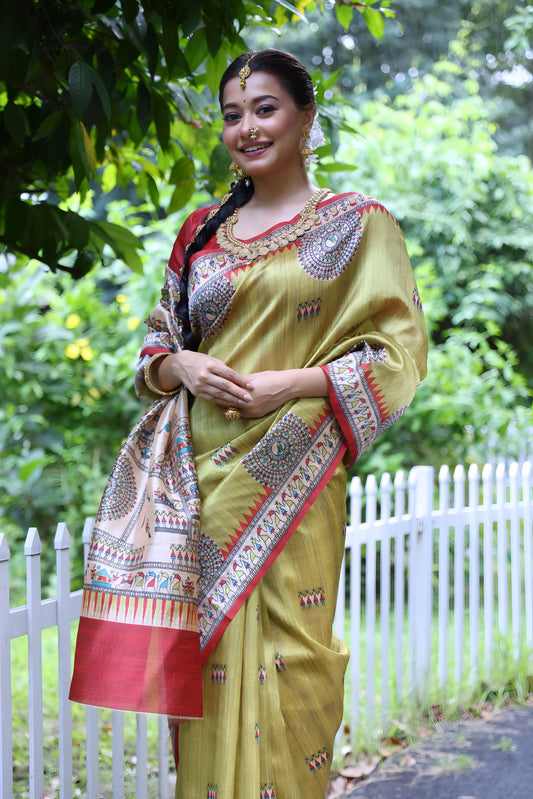 Woman wearing an olive green Madhubani-printed saree with a vibrant red border and intricate traditional motifs, paired with a matching blouse and long braid, elegantly standing on a garden path lined with a white fence and surrounded by lush green foliage.