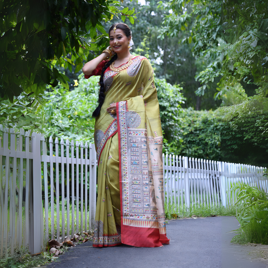 Woman wearing an olive green Madhubani-printed saree with a vibrant red border and intricate traditional motifs, paired with a matching blouse and long braid, elegantly standing on a garden path lined with a white fence and surrounded by lush green foliage.