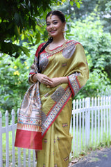 Woman wearing an olive green Madhubani-printed saree with a vibrant red border and intricate traditional motifs, paired with a matching blouse and long braid, elegantly standing on a garden path lined with a white fence and surrounded by lush green foliage.