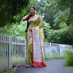 Woman wearing an olive green Madhubani-printed saree with a vibrant red border and intricate traditional motifs, paired with a matching blouse and long braid, elegantly standing on a garden path lined with a white fence and surrounded by lush green foliage.