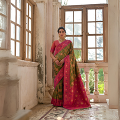 Woman wearing an olive green silk saree with intricate gold floral motifs and a vibrant pink zari border, paired with a matching pink blouse and traditional gold jewelry, standing in a sunlit vintage room with antique wooden windows and ornate architectural details.