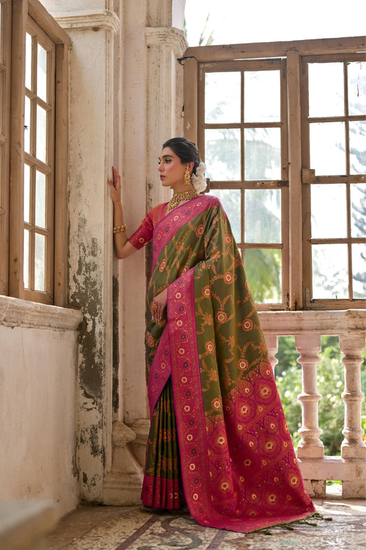 Woman wearing an olive green silk saree with intricate gold floral motifs and a vibrant pink zari border, paired with a matching pink blouse and traditional gold jewelry, standing in a sunlit vintage room with antique wooden windows and ornate architectural details.