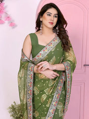Woman wearing an olive green printed saree with traditional yellow motifs and a colorful embroidered border, paired with a matching blouse. She poses gracefully in front of a pastel pink arched doorway, accessorized with silver jewelry, with clay pots, a green plant in a jute vase, and decorative pink floral branches in a modern minimalist studio setting.