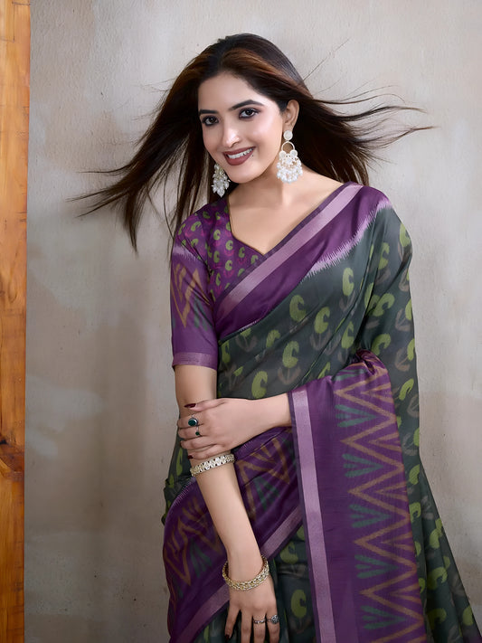  Woman wearing an olive green saree adorned with purple geometric patterns and bold abstract motifs, paired with a matching blouse and statement silver earrings. She poses elegantly in a contemporary studio setting against a neutral textured wall, with a tall woven screen and rustic wooden flooring, showcasing the saree’s vibrant pallu and intricate design.