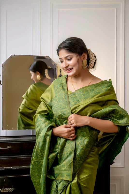 Woman wearing an olive green silk saree with subtle woven patterns, elegantly posing beside a vintage black dresser with a mirror in a softly lit studio featuring white paneled walls and indoor plants.