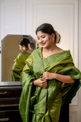 Woman wearing an olive green silk saree with subtle woven patterns, elegantly posing beside a vintage black dresser with a mirror in a softly lit studio featuring white paneled walls and indoor plants.