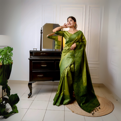 Woman wearing an olive green silk saree with subtle woven patterns, elegantly posing beside a vintage black dresser with a mirror in a softly lit studio featuring white paneled walls and indoor plants.