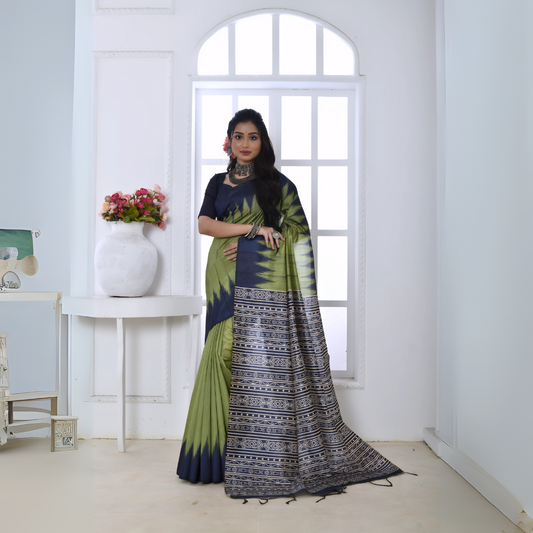 Woman wearing an olive green and navy blue Ikat printed saree with geometric border and pallu, paired with a navy blouse and statement jewelry, standing in a modern white interior with a large arched window and a white console table decorated with a vase of pink flowers.