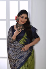 Woman wearing an olive green and navy blue Ikat printed saree with geometric border and pallu, paired with a navy blouse and statement jewelry, standing in a modern white interior with a large arched window and a white console table decorated with a vase of pink flowers.