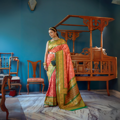 Woman wearing an orange and green silk saree with a gold zari border and intricate patterns, paired with traditional jewelry, standing in a vintage room with blue walls, antique wooden furniture, and a marble floor.