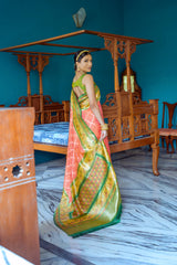 Woman wearing an orange and green silk saree with a gold zari border and intricate patterns, paired with traditional jewelry, standing in a vintage room with blue walls, antique wooden furniture, and a marble floor.