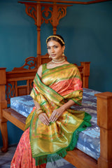 Woman wearing an orange and green silk saree with a gold zari border and intricate patterns, paired with traditional jewelry, standing in a vintage room with blue walls, antique wooden furniture, and a marble floor.