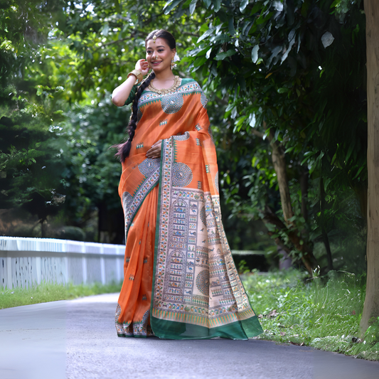 Woman wearing an orange saree with intricate Madhubani-inspired traditional prints and a green border, accessorized with jewelry and a long braid, standing gracefully on a garden path surrounded by lush greenery and trees.