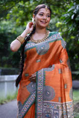 Woman wearing an orange saree with intricate Madhubani-inspired traditional prints and a green border, accessorized with jewelry and a long braid, standing gracefully on a garden path surrounded by lush greenery and trees.