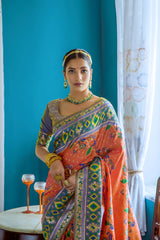 Woman wearing a vibrant orange Patola silk saree with multicolor geometric and floral patterns, paired with a matching blouse and traditional jewelry, elegantly posing in a vintage-inspired room featuring teal blue walls, large windows with white curtains, wooden furniture, and floral decor.