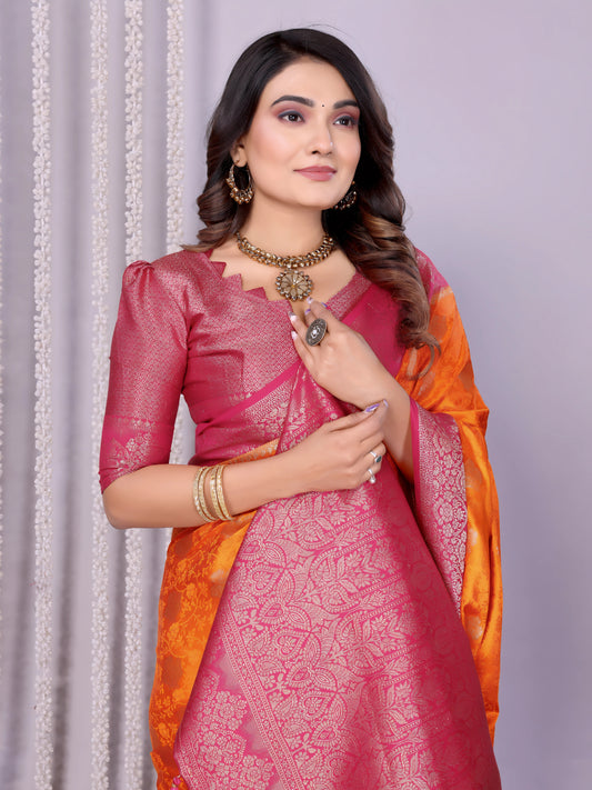Woman wearing a vibrant orange silk saree with a contrasting pink blouse and ornate golden border, featuring traditional woven motifs and an intricate pallu design. She is styled with gold jewelry and bangles, posing gracefully in a modern studio adorned with decorative flower baskets, a neutral backdrop, and subtle floral accents.