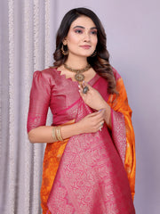 Woman wearing a vibrant orange silk saree with a contrasting pink blouse and ornate golden border, featuring traditional woven motifs and an intricate pallu design. She is styled with gold jewelry and bangles, posing gracefully in a modern studio adorned with decorative flower baskets, a neutral backdrop, and subtle floral accents.
