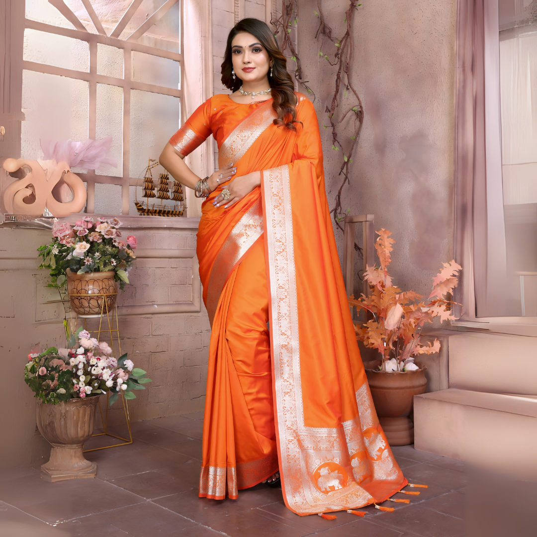 Woman wearing a vibrant orange silk saree with a contrasting gold zari border and pallu featuring traditional motifs, paired with a matching blouse. She is styled with silver jewelry and poses gracefully in a vintage-inspired floral studio with pastel blossoms, decorative vases, and a large window in the background.
