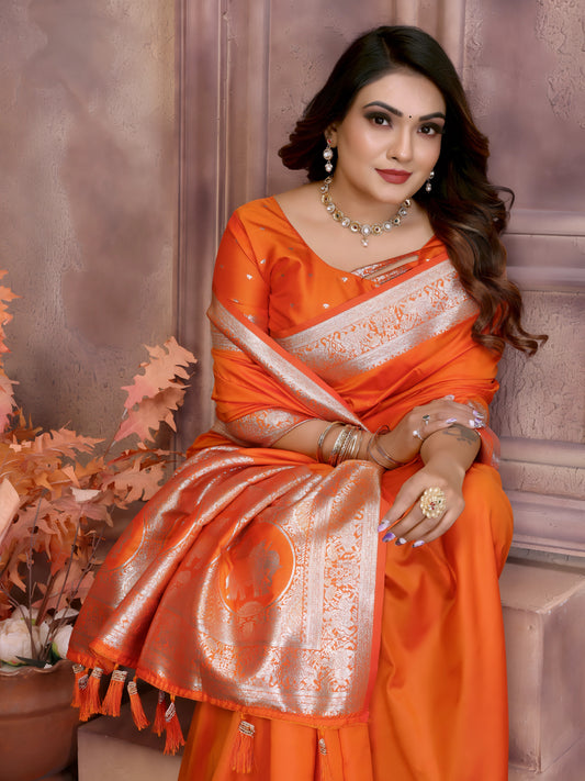 Woman wearing a vibrant orange silk saree with a contrasting gold zari border and pallu featuring traditional motifs, paired with a matching blouse. She is styled with silver jewelry and poses gracefully in a vintage-inspired floral studio with pastel blossoms, decorative vases, and a large window in the background.