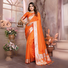 Woman wearing a vibrant orange silk saree with a contrasting gold zari border and pallu featuring traditional motifs, paired with a matching blouse. She is styled with silver jewelry and poses gracefully in a vintage-inspired floral studio with pastel blossoms, decorative vases, and a large window in the background.