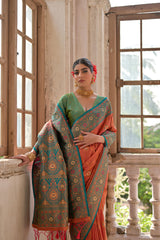 Woman wearing an orange silk saree with intricate gold and turquoise border, paired with a green blouse and traditional jewelry, standing by antique wooden windows in a vintage architectural setting with natural light streaming in.