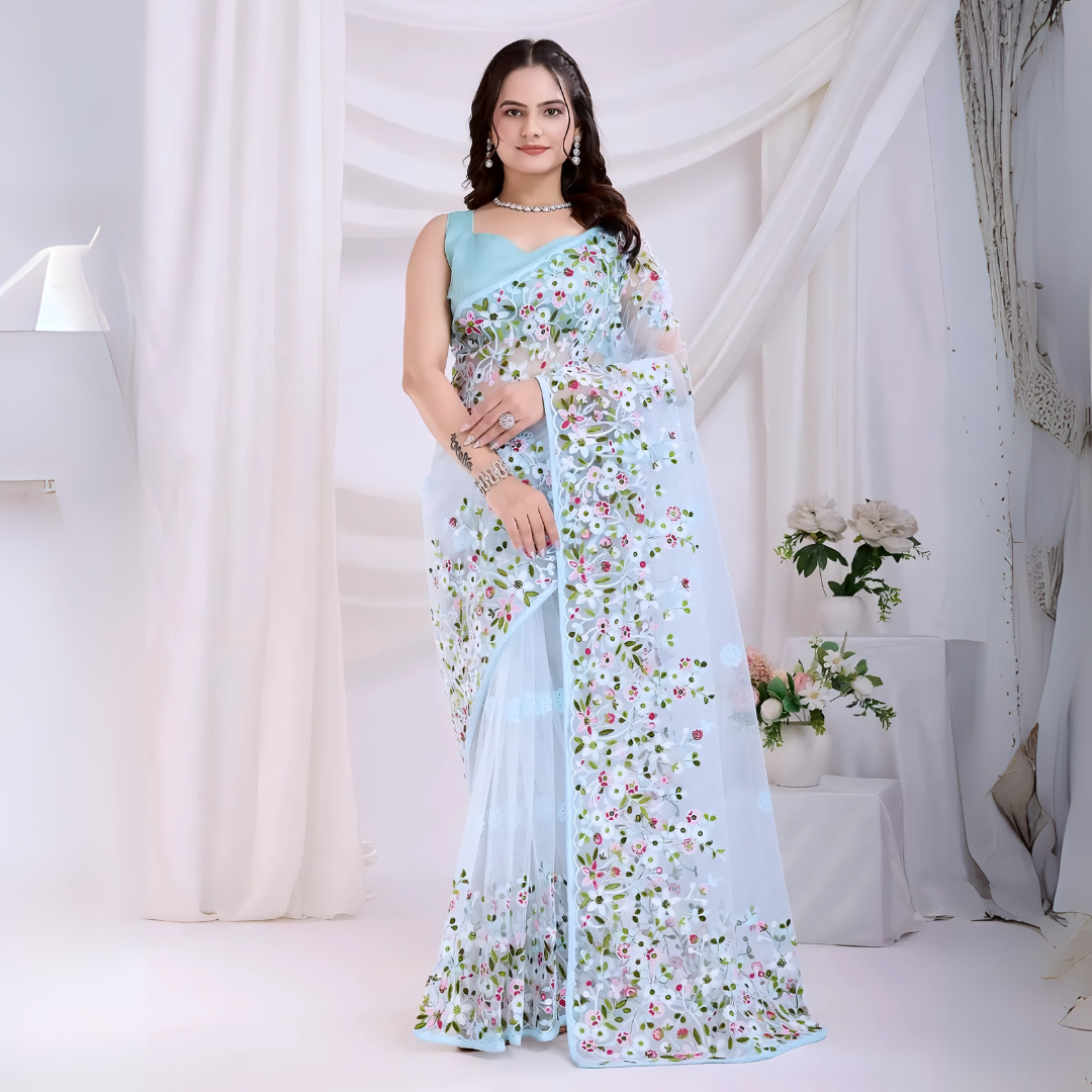 Woman wearing a pastel blue net saree with intricate multicolor floral embroidery and a matching blue sleeveless blouse, posing in a modern white studio with draped curtains and floral arrangements