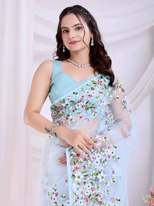 Woman wearing a pastel blue net saree with intricate multicolor floral embroidery and a matching blue sleeveless blouse, posing in a modern white studio with draped curtains and floral arrangements