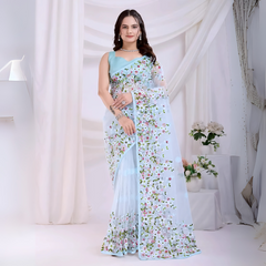 Woman wearing a pastel blue net saree with intricate multicolor floral embroidery and a matching blue sleeveless blouse, posing in a modern white studio with draped curtains and floral arrangements