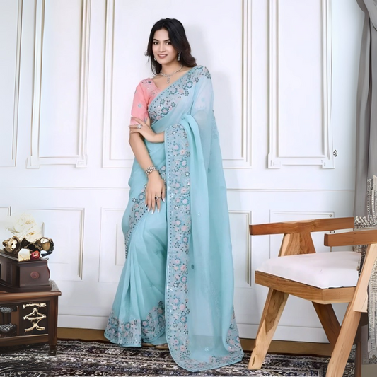  Woman wearing a pastel blue saree with intricate floral embroidery on the border, paired with a pink embroidered blouse, posing in a modern white interior with a wooden chair, decorative table, and floral arrangement.