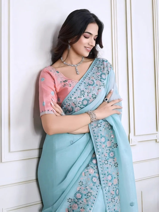  Woman wearing a pastel blue saree with intricate floral embroidery on the border, paired with a pink embroidered blouse, posing in a modern white interior with a wooden chair, decorative table, and floral arrangement.