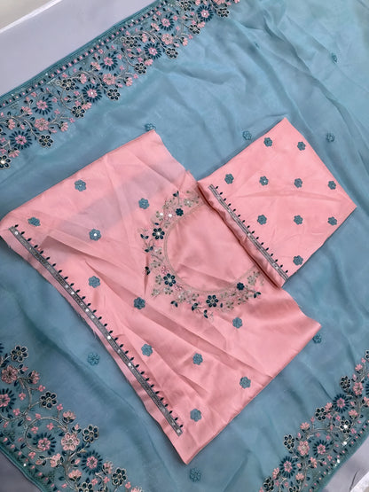  Woman wearing a pastel blue saree with intricate floral embroidery on the border, paired with a pink embroidered blouse, posing in a modern white interior with a wooden chair, decorative table, and floral arrangement.