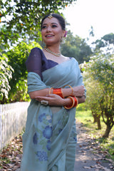 Woman wearing a pastel blue silk saree with subtle floral embroidery and a navy blue blouse, accessorized with traditional jewelry and orange bangles, standing beside a white picket fence in a sunlit garden surrounded by lush green trees.