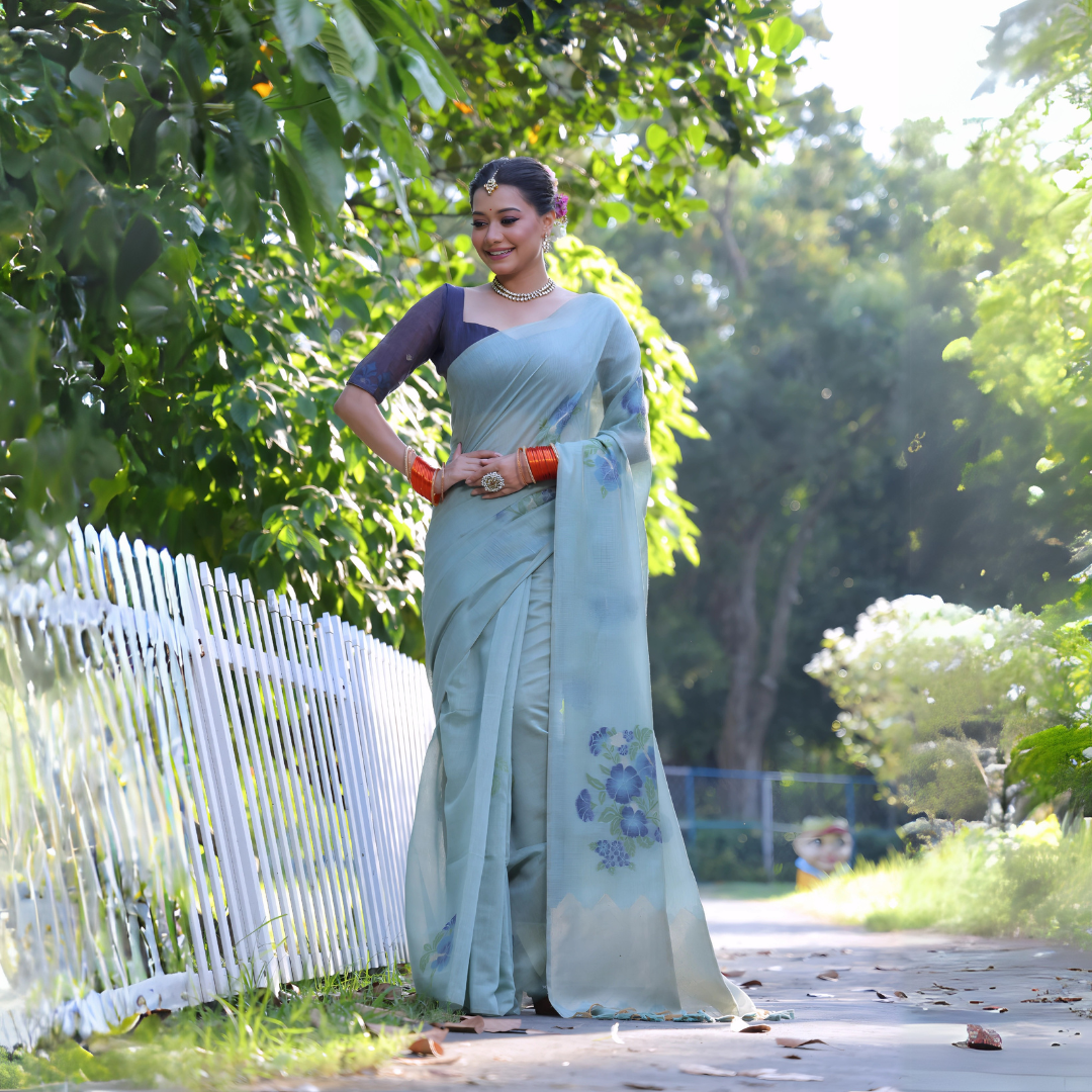 Woman wearing a pastel blue silk saree with subtle floral embroidery and a navy blue blouse, accessorized with traditional jewelry and orange bangles, standing beside a white picket fence in a sunlit garden surrounded by lush green trees.