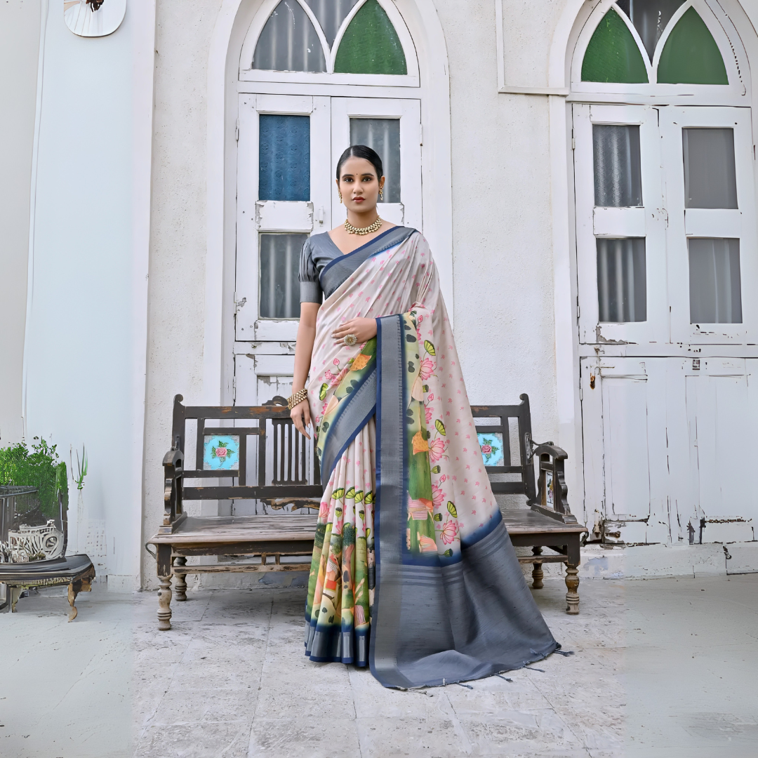 Woman wearing a pastel floral silk saree with a grey border and matching grey blouse, accessorized with traditional gold jewelry, elegantly standing in front of classic white arched doors and windows beside a vintage wooden bench, with greenery and a floral arrangement in a heritage courtyard setting.