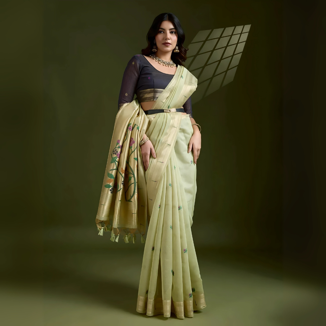Woman wearing a pastel green silk saree with subtle embroidered motifs and golden zari border, paired with a black three-quarter sleeve blouse and traditional jewelry, elegantly posing in a contemporary studio with olive green walls and artistic window light patterns.