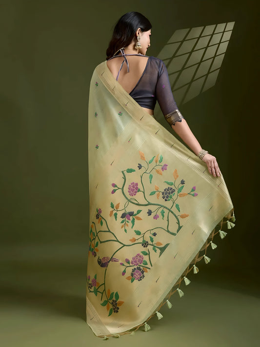 Woman wearing a pastel green silk saree with subtle embroidered motifs and golden zari border, paired with a black three-quarter sleeve blouse and traditional jewelry, elegantly posing in a contemporary studio with olive green walls and artistic window light patterns.