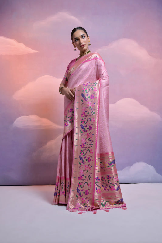Woman wearing a pastel pink Banarasi silk saree with an intricate multicolor floral zari border and pallu, paired with a matching pink blouse and traditional jewelry, posing elegantly in a pastel-themed studio with dreamy cloud backgrounds