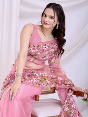 Woman wearing a pastel pink net saree with intricate white and green floral embroidery and a matching sleeveless blouse, posing in a modern white studio with draped curtains and floral arrangements.