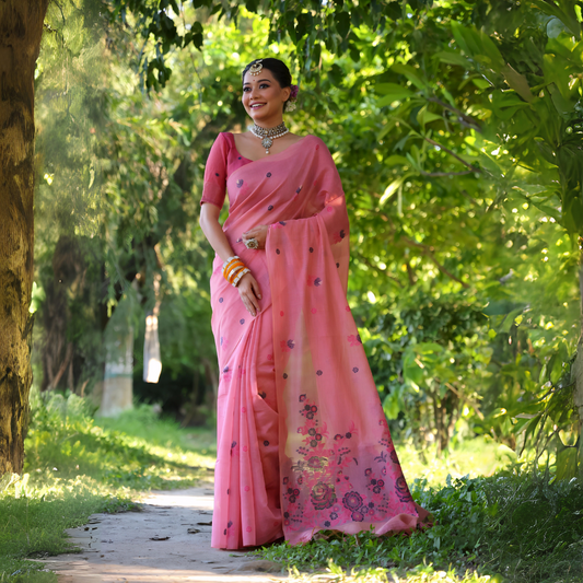 Woman wearing a pastel pink saree with delicate floral embroidery and a matching pink blouse, accessorized with silver jewelry and colorful bangles, standing on a sunlit garden path surrounded by lush green trees and foliage.