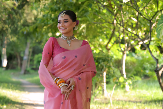 Woman wearing a pastel pink saree with delicate floral embroidery and a matching pink blouse, accessorized with silver jewelry and colorful bangles, standing on a sunlit garden path surrounded by lush green trees and foliage