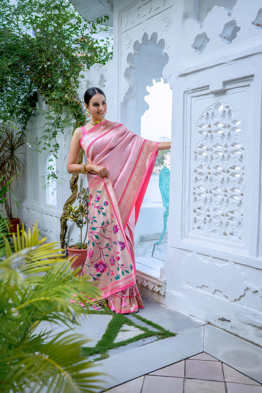 Woman wearing a pastel pink silk saree with gold border and handpainted floral pallu, styled with a matching blouse and gold jewelry, posing in a white heritage courtyard surrounded by green potted plants and traditional architectural details