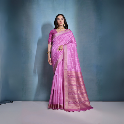  Woman wearing a pastel pink silk saree with subtle woven motifs and a gold zari border, paired with a matching blouse and traditional gold jewelry, posing against a soft blue gradient studio background.