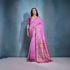  Woman wearing a pastel pink silk saree with subtle woven motifs and a gold zari border, paired with a matching blouse and traditional gold jewelry, posing against a soft blue gradient studio background.