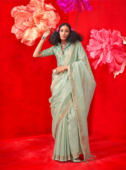 A person wearing a pastel sea green saree with intricate zari work, accessorized with bangles and a necklace, standing against a vibrant red background with large, colorful flowers.