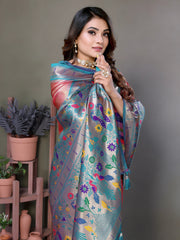 Woman wearing an elegant peach pink silk saree with a striking blue and silver zari border and intricate multicolor floral motifs on the pallu, paired with a blue and purple blouse. She is accessorized with traditional gold jewelry, bangles, and rings, posing gracefully in a contemporary studio featuring potted green plants and terracotta vases on pastel wooden stands against a neutral backdrop.