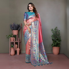 Woman wearing an elegant peach pink silk saree with a striking blue and silver zari border and intricate multicolor floral motifs on the pallu, paired with a blue and purple blouse. She is accessorized with traditional gold jewelry, bangles, and rings, posing gracefully in a contemporary studio featuring potted green plants and terracotta vases on pastel wooden stands against a neutral backdrop.