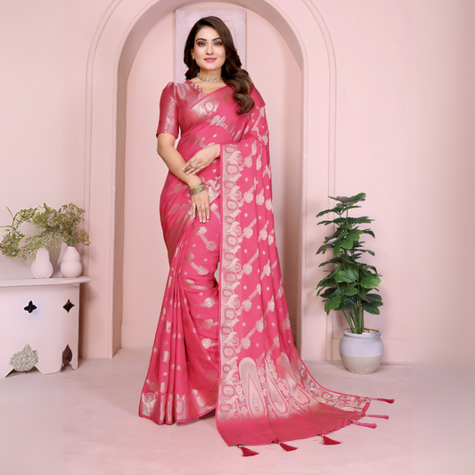 Woman wearing a pink Banarasi saree featuring intricate gold zari motifs and a decorative border with tassel details, paired with a matching blouse with short sleeves. She stands gracefully in front of a pastel arched backdrop, styled with traditional jewelry and bangles, with minimal decor including a white pedestal with vases and a potted green plant, showcasing a festive ethnic look.